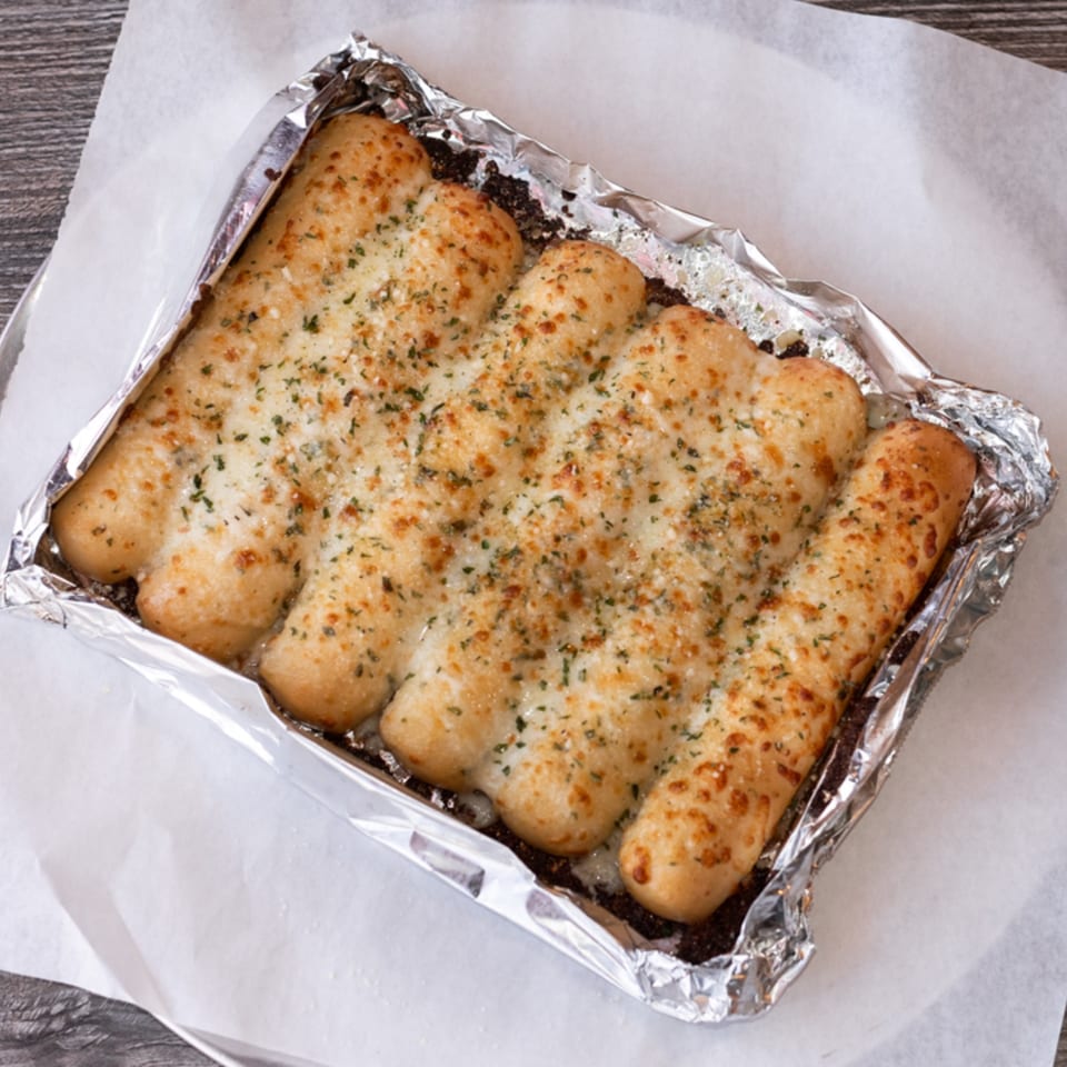 Golden-brown garlic breadsticks, topped with herbs and cheese, in a foil-lined container.