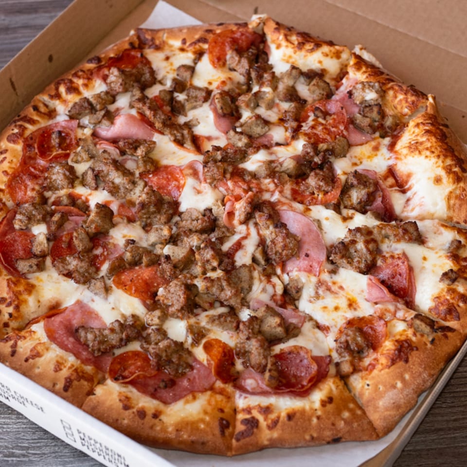 A Fiesta Pizza meat lover's pizza, topped with pepperoni, ham, and sausage, in a cardboard box.