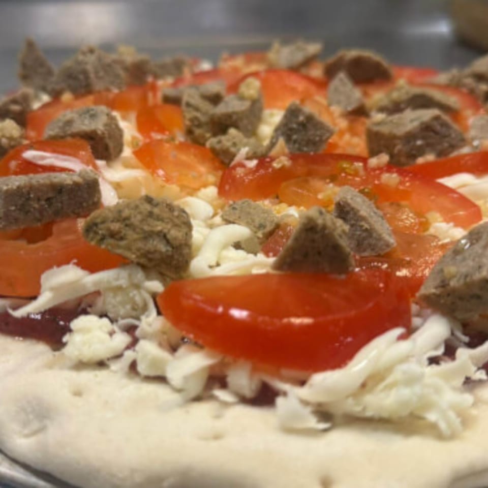 Uncooked Fiesta Pizza with tomato slices, cheese, and sausage crumbles on a dough base.